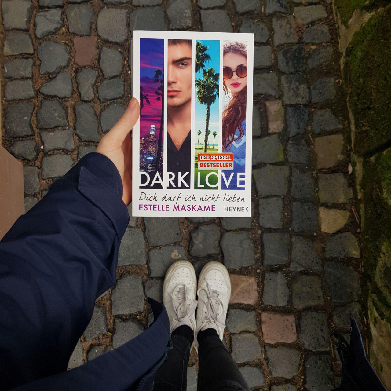 Foto: Eine Hand hält das Buch von Estelle Maskame "Dark Love". Im Hintergrund sieht man weiße Schuhe und den Gehweg.