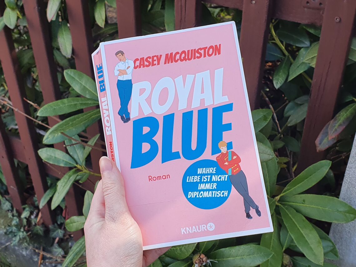 Hand hält Buch. Buch hat ein Rosa Cover und den Titel "Royal Blue". Außerdem sind 2 Männer auf dem Cover.