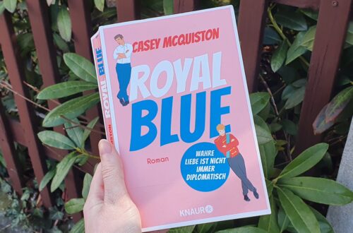 Hand hält Buch. Buch hat ein Rosa Cover und den Titel "Royal Blue". Außerdem sind 2 Männer auf dem Cover.